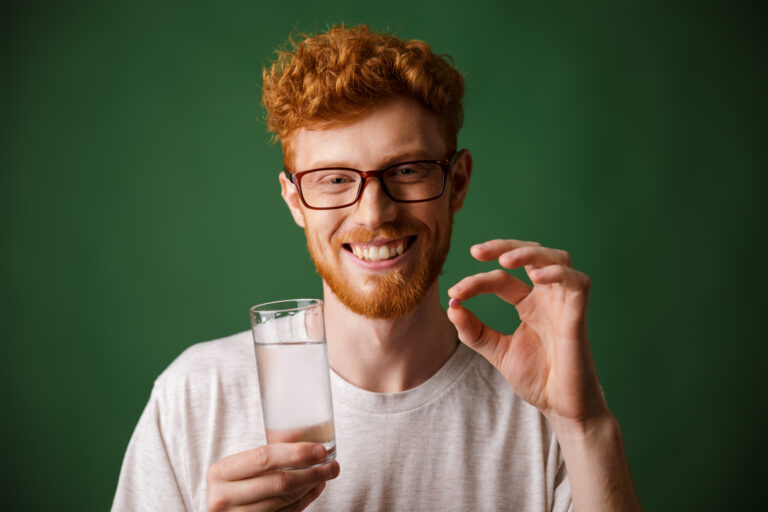 Man with red hair smiling, holding a finasteride tablet and a glass of water
